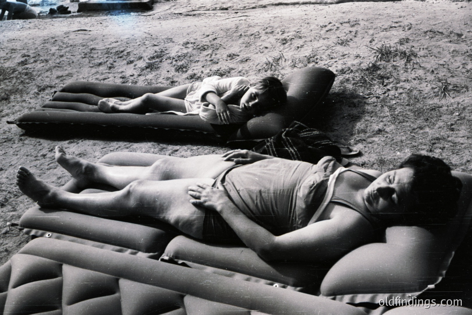 Two young women recline on inflatable mattresses in a grassy, outdoor setting, likely a backyard. They appear relaxed, some wearing casual clothing. The grainy, black-and-white aesthetic suggests a snapshot from the 1970s. Could serve as reference for vintage lifestyle imagery.
