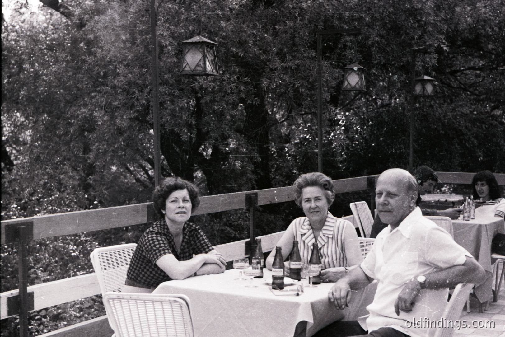 A black and white photograph depicts three people seated at an outdoor table covered with a white cloth. A fourth individual is visible blurred in the background. The setting appears to be a restaurant terrace with foliage and lanterns. Likely 1960s, capturing a moment of leisure.