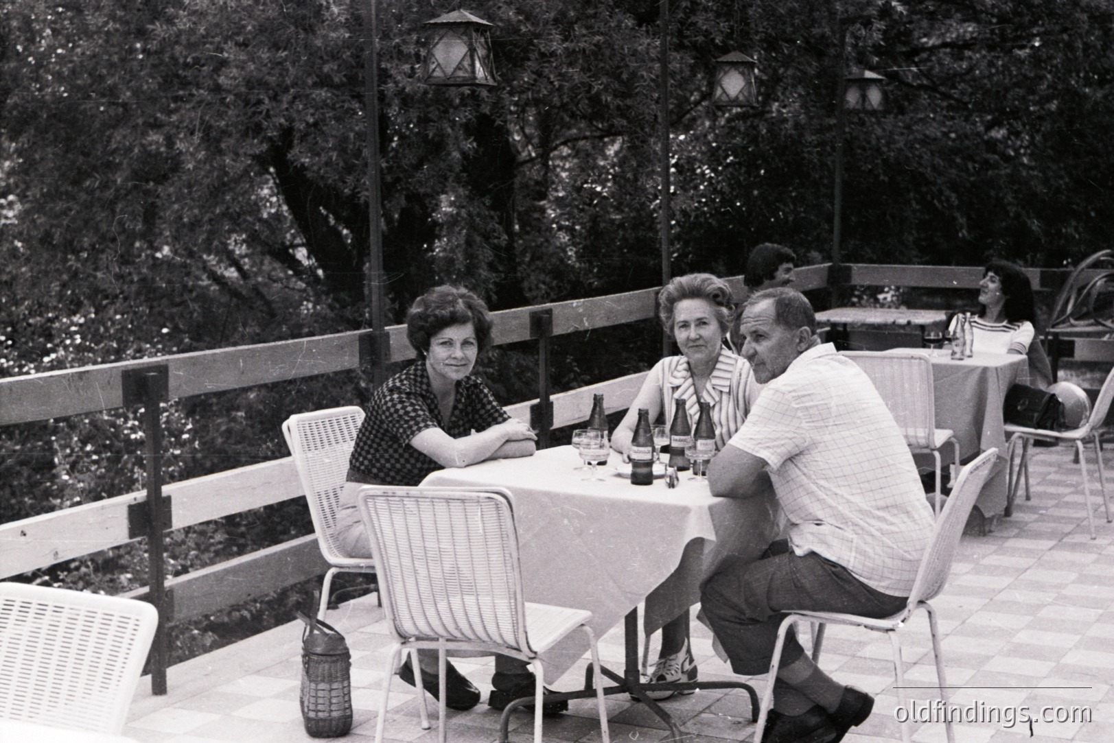 Three people sit at a table covered in a white cloth, enjoying a meal outdoors. A partial view of additional diners is visible in the background. Features of 1960s/70s dress & design evident. Balcony overlooks a dense wooded area. Likely a resort or restaurant setting.