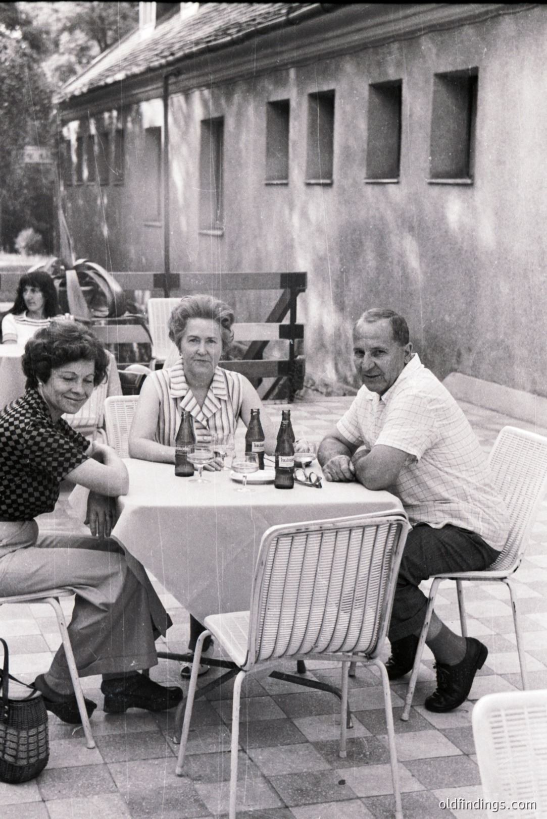 A family gathers around a table setting on a patio, likely in the 1960s. Three adults are seated – a woman in a dark floral top, a matronly figure, and a man in a short-sleeved shirt. Several bottles of beer suggest a casual gathering.