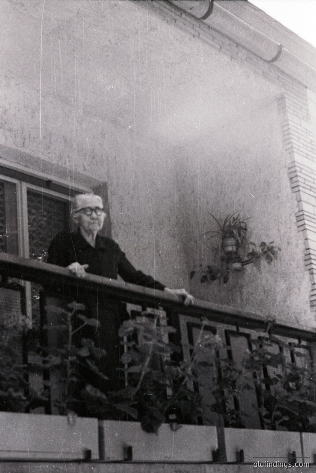 An elderly man stands on a balcony, appearing contemplative. Architectural details suggest a mid-century modern building with exposed brick. Balcony features decorative railing and planters with greenery. Likely taken in the 1960s or 70s. A wall-mounted lamp illuminates the scene.