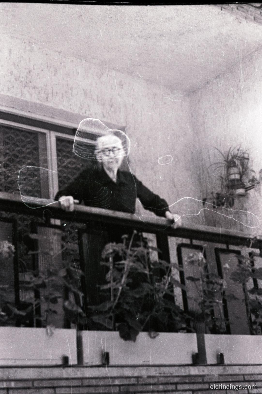 A monochrome image captures an older woman on a balcony. She wears a dark, buttoned shirt and stands with hands on the railing. Lush foliage fills planters below. The balcony's brick base and railing suggest a mid-century architectural style. Likely 1960s-1970s. A sense of quietude and domesticity. Suitable for architectural or lifestyle stock.