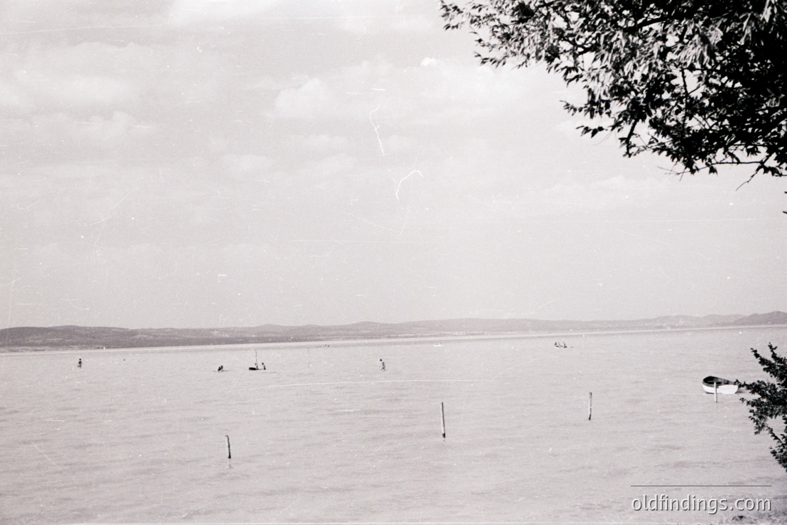 A wide coastal view shows figures wading and boating in calm water, with a low, distant shoreline visible through a hazy atmosphere. Several wooden stakes protrude from the water. Likely a seaside resort or fishing area. Appears to be a mid-20th century snapshot.