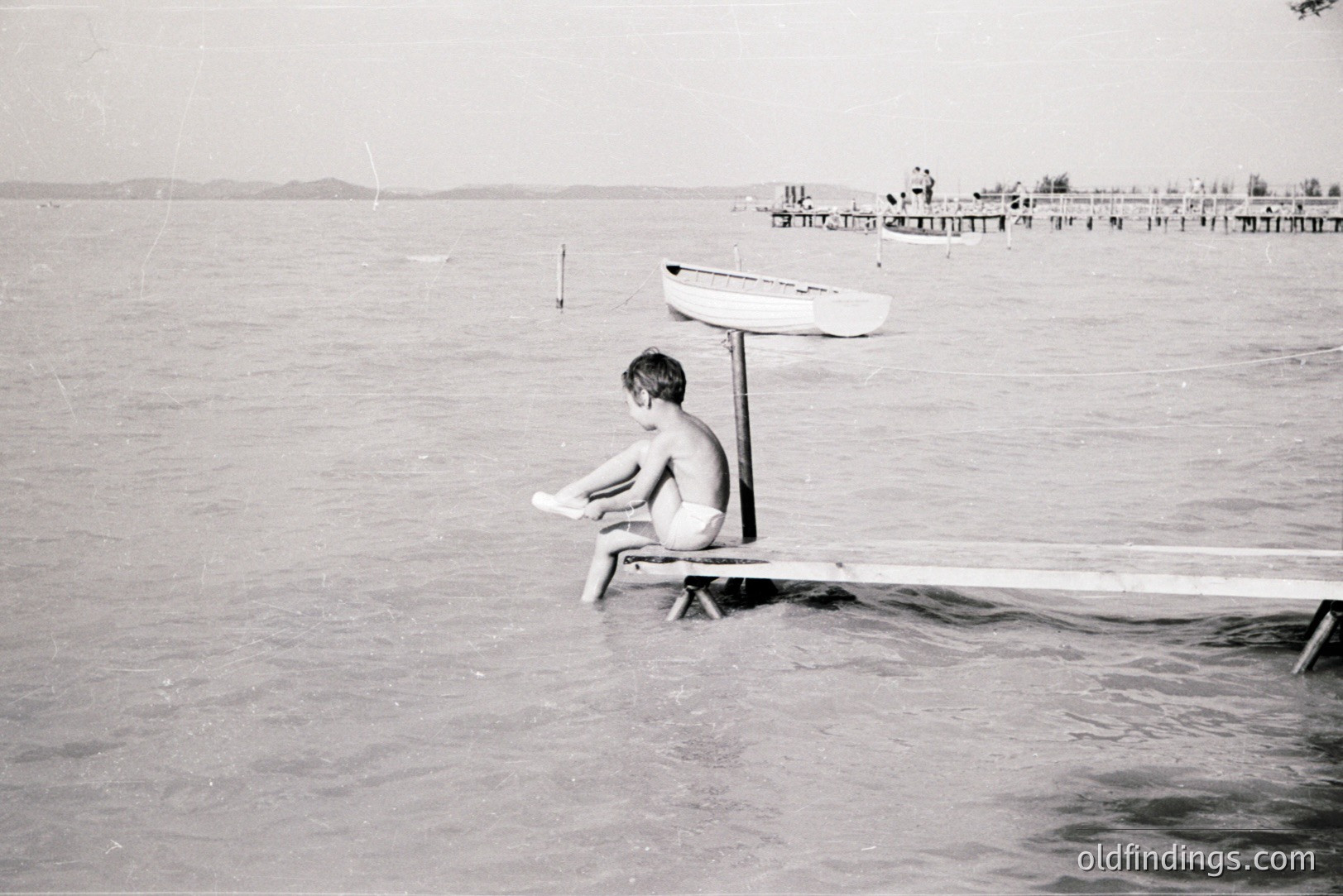 A young boy sits pensively on a pier, partially submerged in water. A small boat is secured overhead. Visible in the distance is a crowded pier with onlookers and several small boats. Appears to be a seaside resort scene. Likely 1960s, possibly Eastern Europe.
