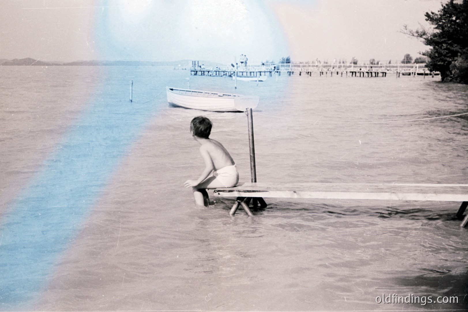 A young boy, viewed from behind, sits on a weathered wooden pier extending into a tranquil lake. A small sailboat rests near the pier. The shoreline features a wooden structure with what appear to be buildings. The photo’s aesthetic suggests a mid-20th century, possibly 1950s, snapshot.