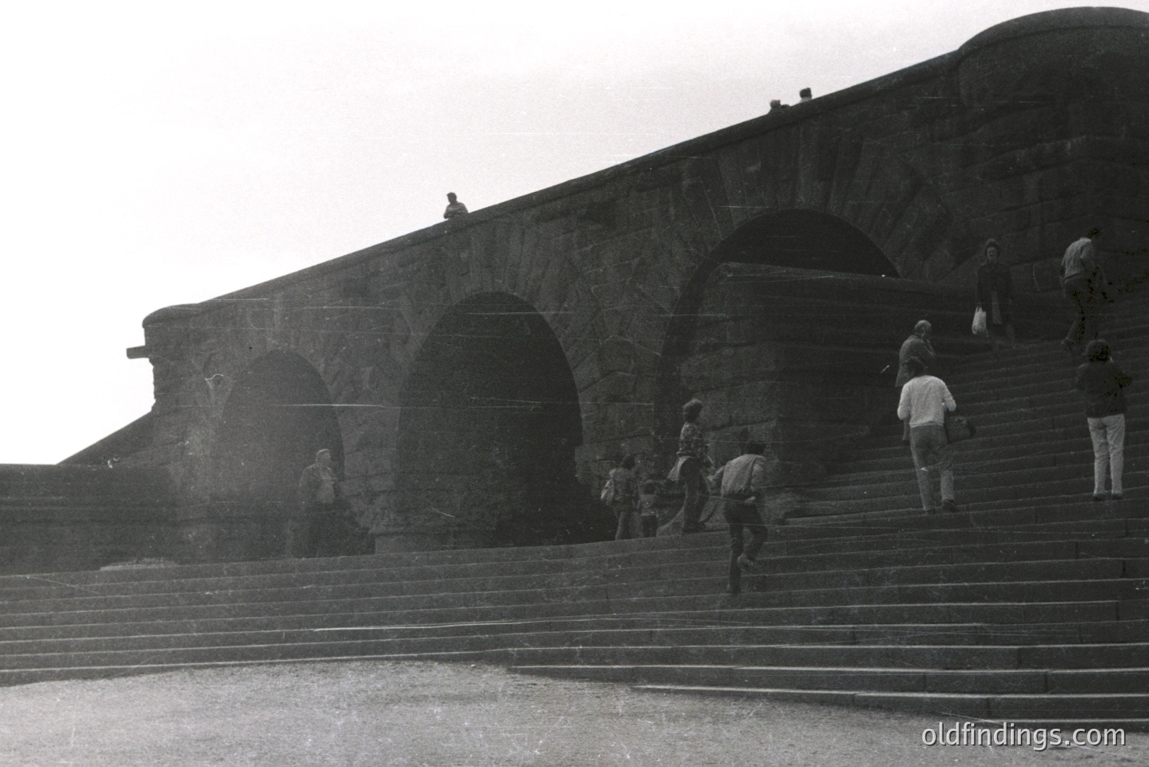 Striking black & white image of tiered stone steps ascending to a monumental, arched structure, possibly a fortress or public building. Several figures are climbing the steps, while others are visible in the archways. Architecture suggests late 1960s or 1970s. Likely seaside location.