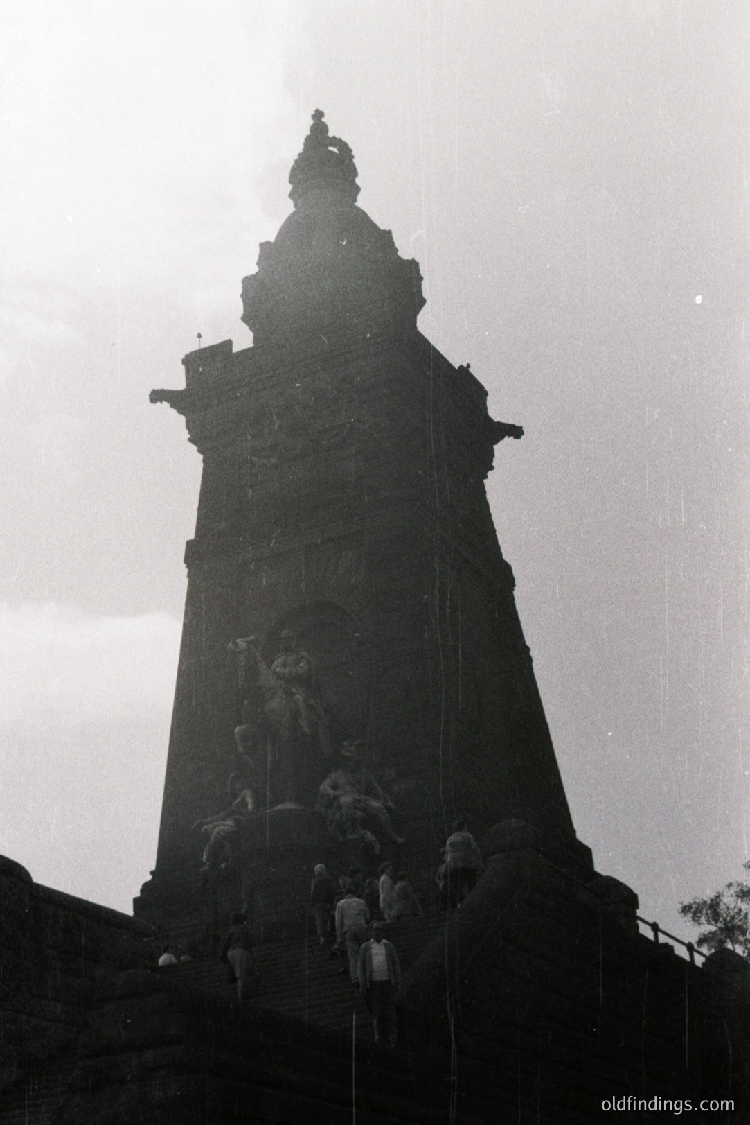 Monumental stone structure, likely a memorial or triumphal arch, rises dramatically against a clouded sky. A small group of people ascends a stone staircase leading to the base. Appears to be a low-angle shot emphasizing scale. Likely mid-20th century, potentially post-war era.