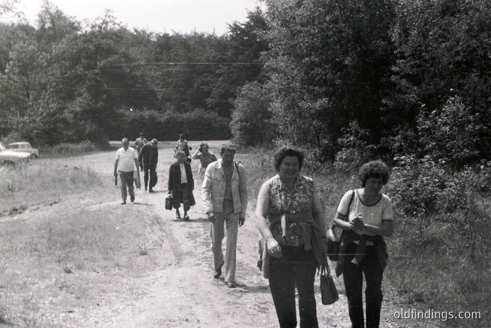 Group of people walk along a rural road, likely in Bulgaria. The image captures casual attire of the 1970s - wide-legged trousers, patterned tops, and short hairstyles. A vintage car is visible in the distance. Likely a snapshot of everyday life.