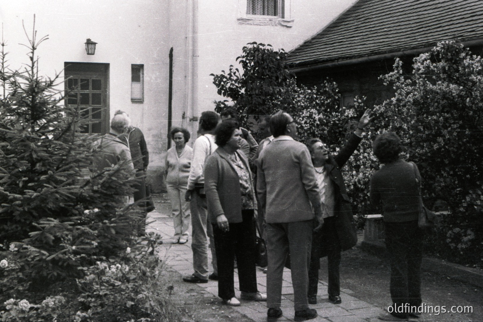 A group of formally-dressed people walk along a stone path toward a building facade. Architectural details show a mix of brick and stucco. The scene is overgrown with foliage and a small evergreen. Likely a courtyard or garden setting, mid-1970s. Suitable for design and historical reference.