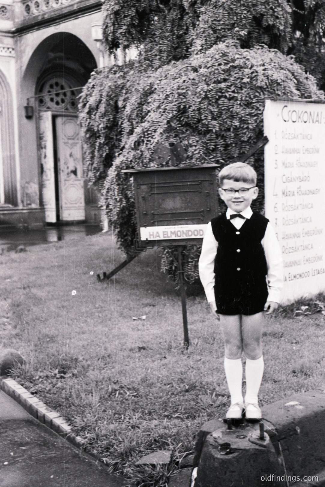 A young boy, likely in the 1960s, stands proudly atop a low stone pedestal. He wears a formal outfit: a bow tie, vest, collared shirt, and shorts, with knee-high socks. A sign reads “HA ELMONDOD” with text below. Overgrown foliage and an arched doorway form the backdrop. The scene evokes a sense of formality and perhaps a theatrical or school-related event.