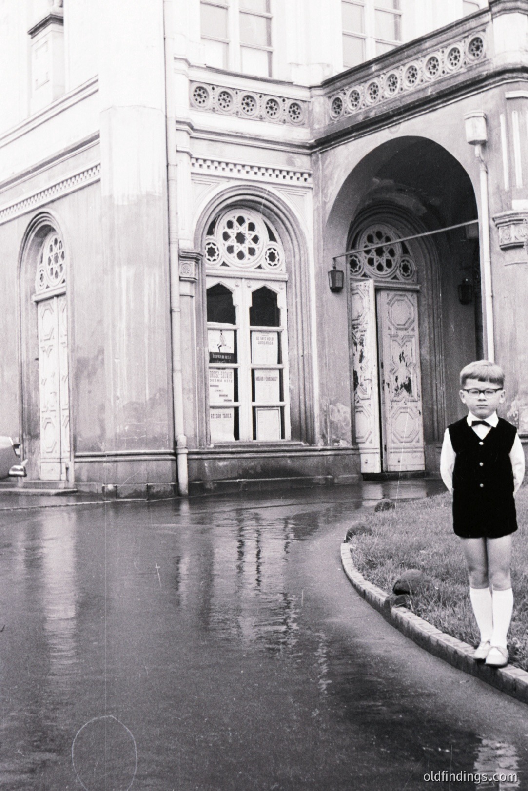 A young boy in a dark vest and knee socks stands on a wet road, with an ornate building facade visible behind him. Architectural details show an arched doorway and decorative stonework. Likely 1960s-70s. Evokes a nostalgic, slightly melancholic mood. Could be used for design or historical research.