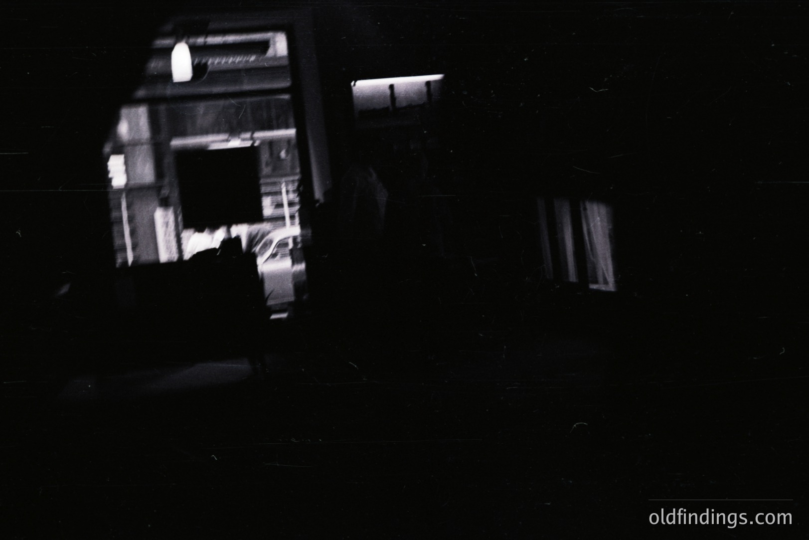 A black and white photograph depicts a car within a garage. Light from the garage spills outward, illuminating a partial view of a vehicle and architectural details. The image has a grainy texture, suggestive of film photography. Likely 1960s-1970s style.