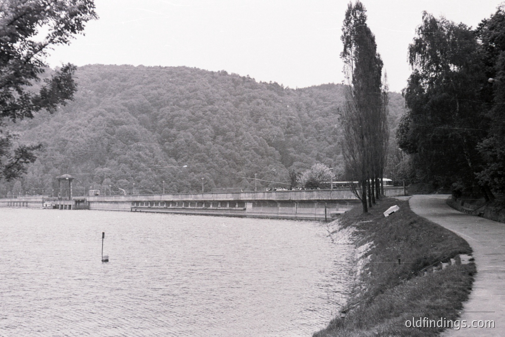 Scenic view of a lakeside structure with a curving pathway along the bank. Lush forested hillside rises dramatically in the background. Appears to be a reservoir or dam, possibly with a control building visible. Likely mid-20th century, based on photographic style.