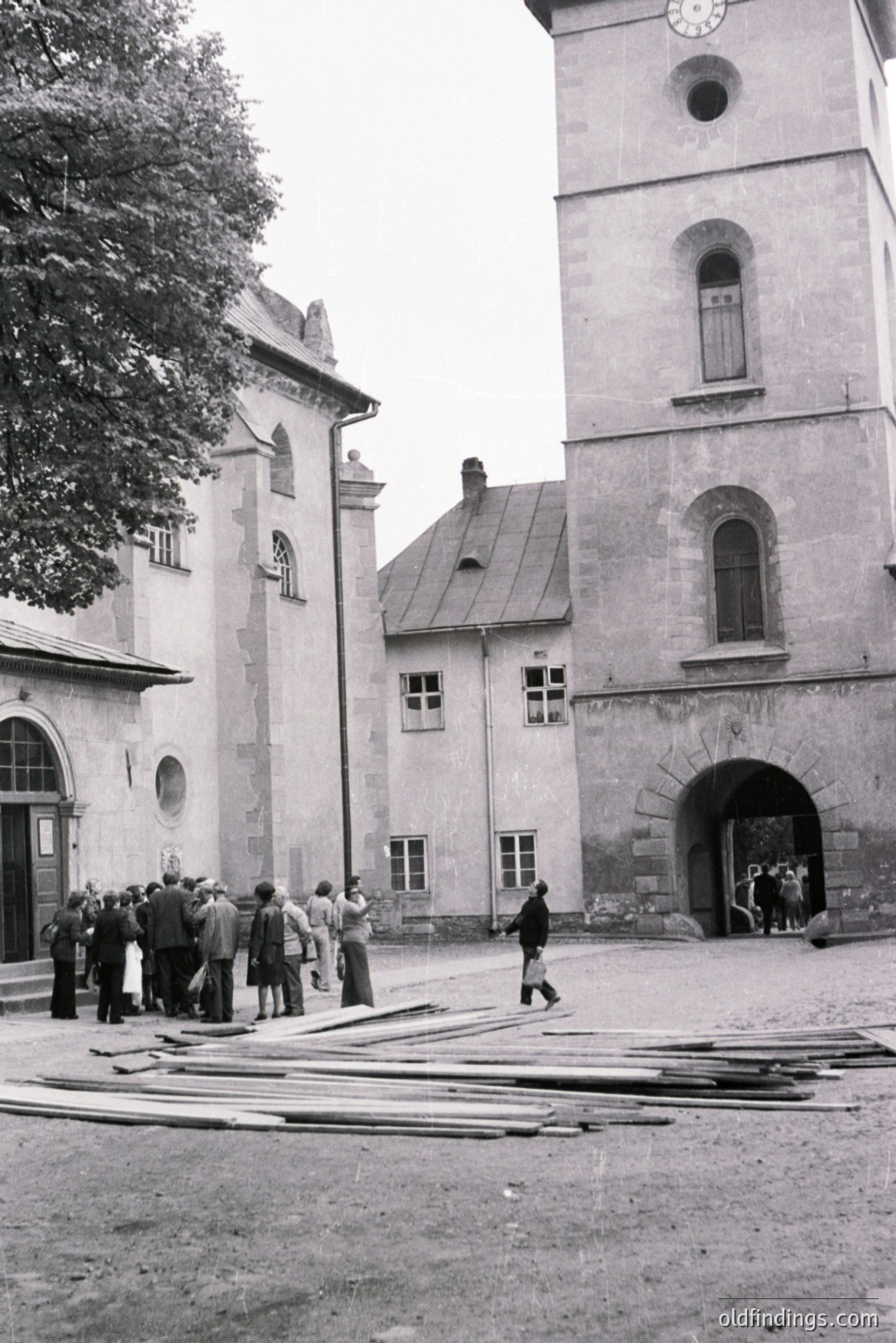 A gray, monochrome image showcases a medieval courtyard with a prominent stone tower featuring arched windows and a clock face. A group of people in period clothing stands near a building with a detailed doorway. Scattered lumber suggests construction or repair work. Likely 1950s-1970s.