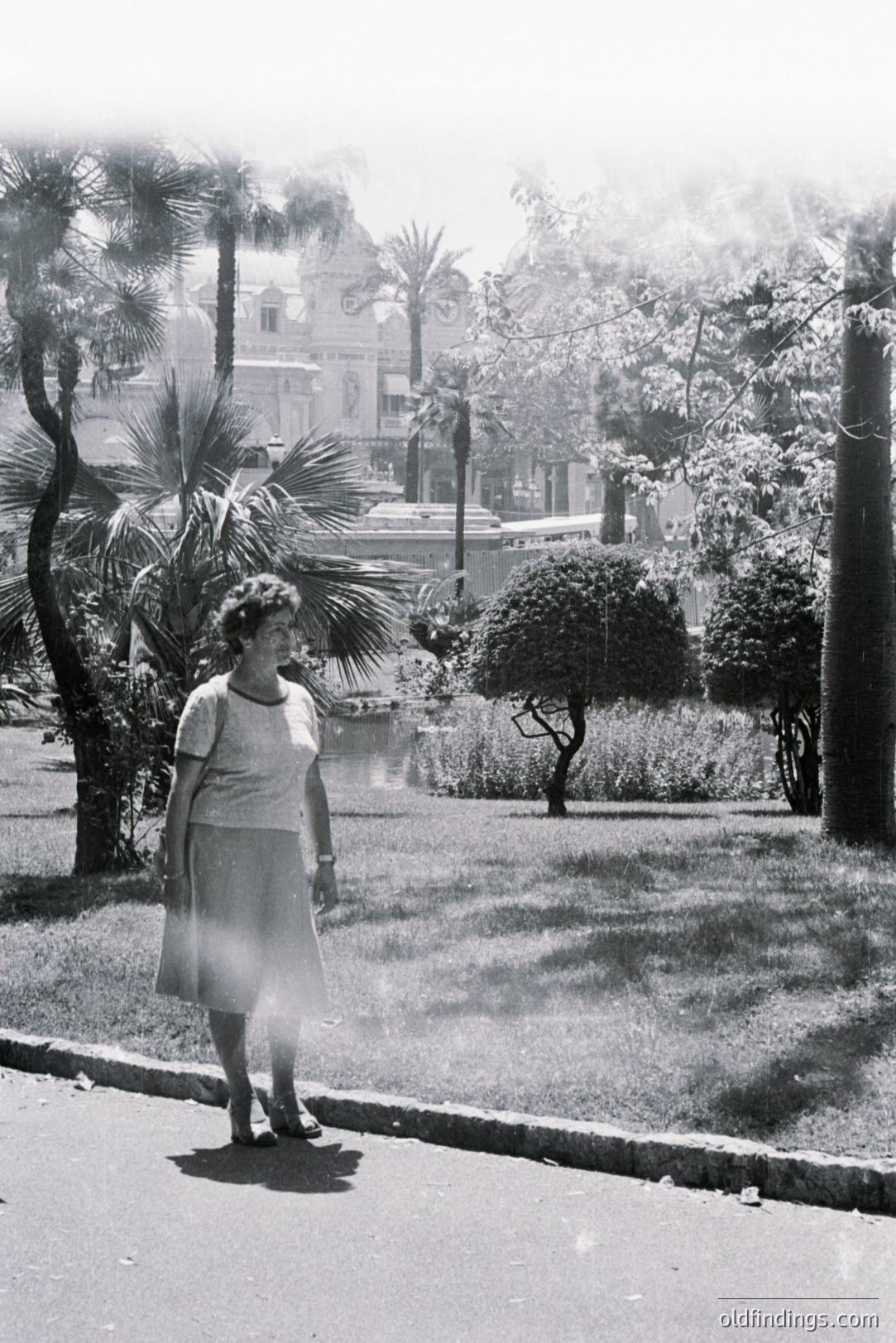 A woman in a simple dress and sandals stands beside a paved road, framed by lush tropical foliage and a waterway. A grand, ornate building is visible across the water. Likely 1960s, suggestive of a seaside resort town. Potential stock image for fashion or travel themes.