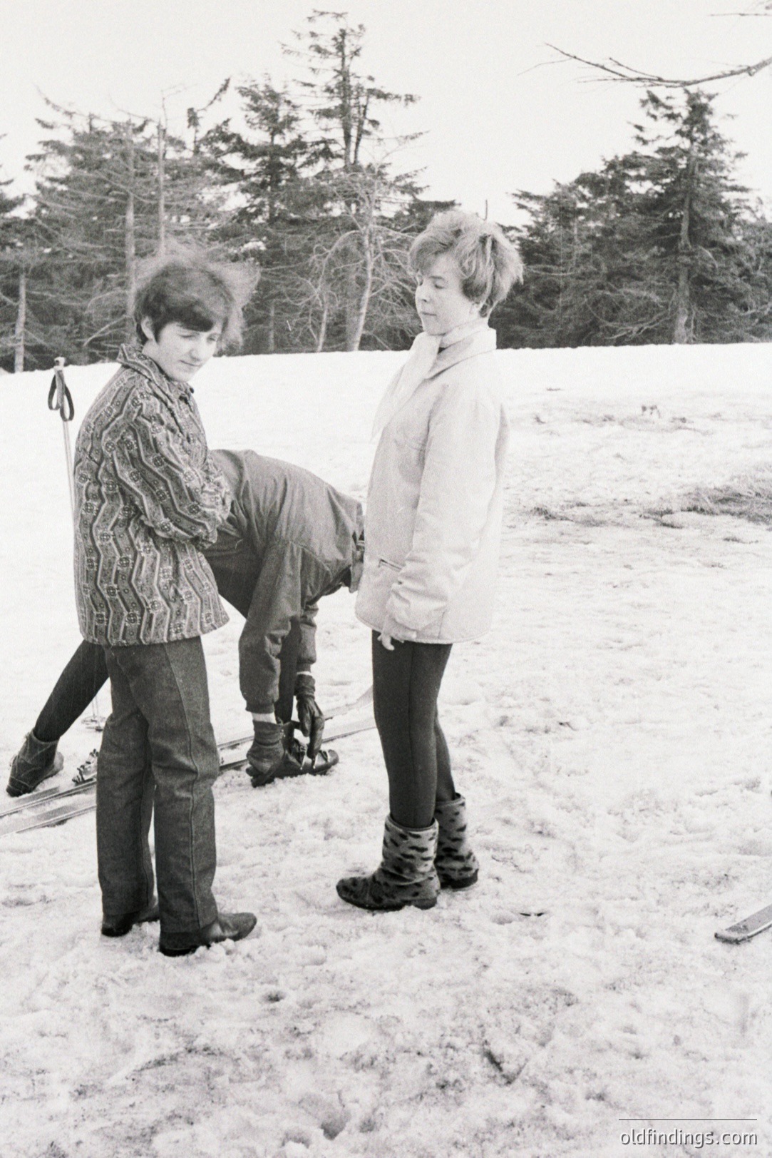 Two individuals, one tangled in skis and fallen in the snow, the other looking on. A wintry landscape with evergreen trees dominates the background. Clothing styles suggest a 1960s-70s era, with corduroy pants and bulky outerwear. Likely a recreational skiing scene.