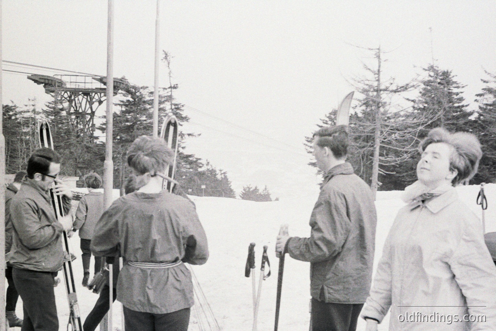 Four figures stand amidst snow-covered evergreens, near a ski lift tower. Two men, one smoking, and two women in 1960s winter attire converse while holding skis. Clothing style suggests a leisure activity. Documentary style, likely a candid snapshot.