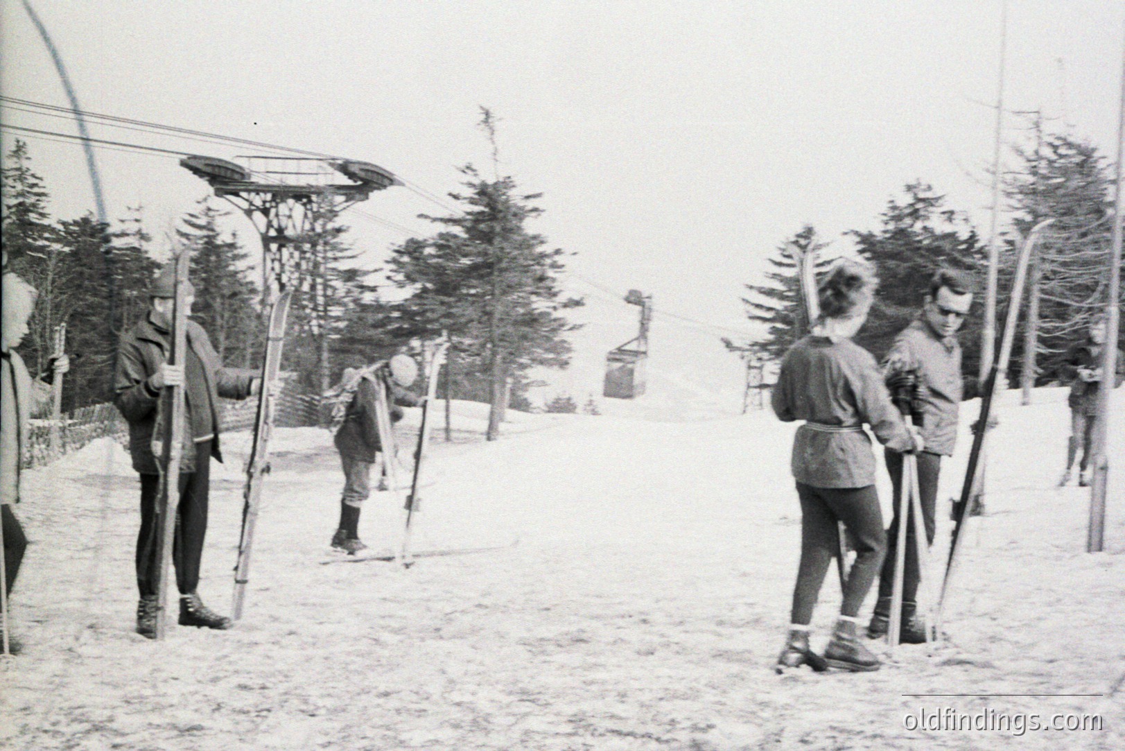 Vintage photo depicts skiers at a rope tow lift, likely in the 1940s or 50s. Several individuals are poised to ride or disembark, bundled in winter clothing. Sparse trees and a snowy landscape contribute to the rural, alpine atmosphere. A functional, utilitarian aesthetic characterizes this scene.