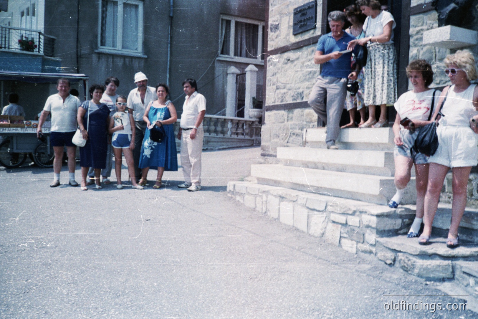 A group gathers on steps outside a stone building; architecture suggests a European coastal setting. Fashion trends indicate the 1970s - wide collars, short hemlines, and oversized sunglasses are visible. A red scooter is parked nearby. Likely a travel snapshot.