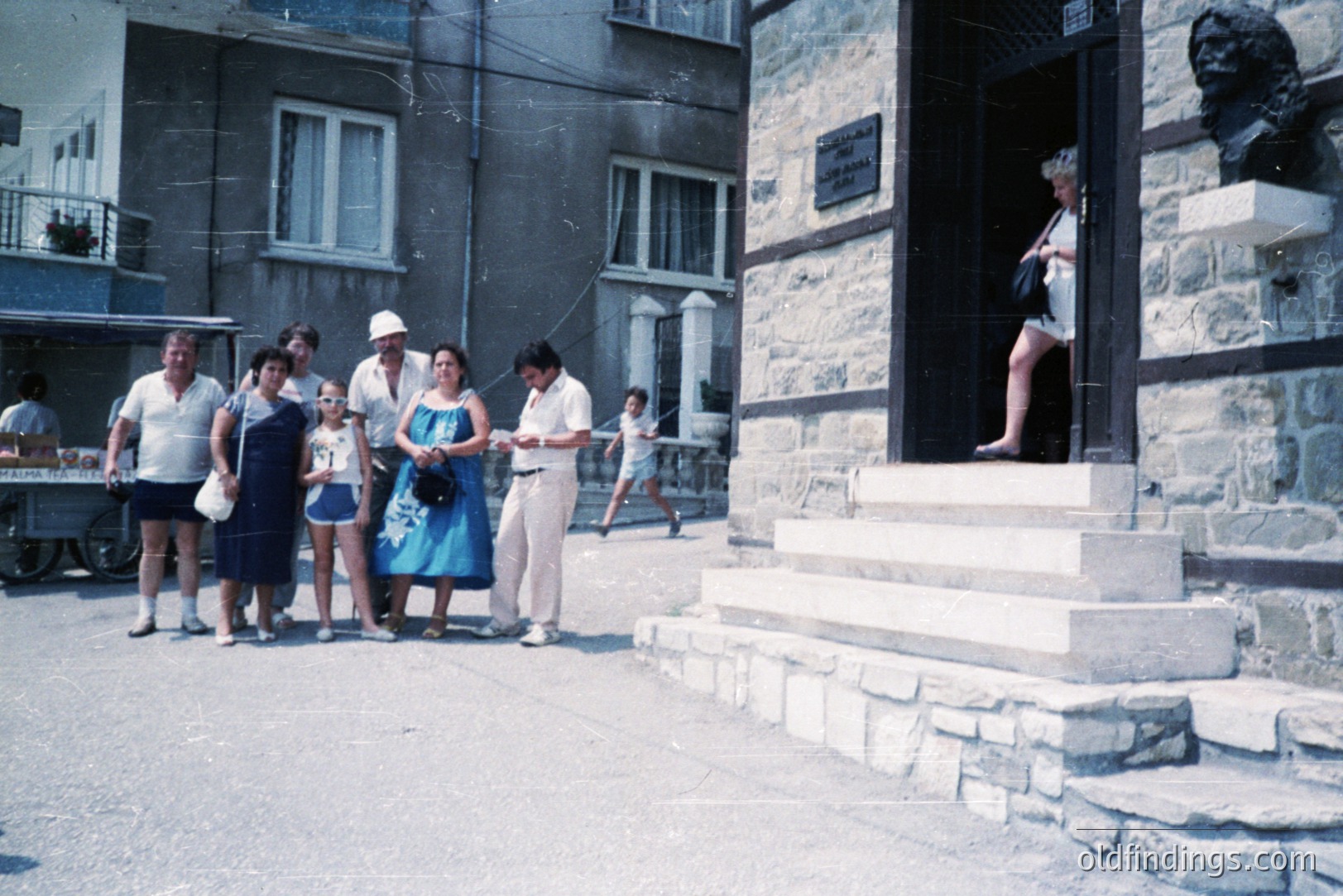 A group of tourists, dressed in 1970s attire, poses outside a stone building. A bust of a man sits atop the building's entry. A young boy runs down the sidewalk alongside. Appears to be a seaside town, likely Eastern Europe. The building's architecture hints at a historical or cultural significance.