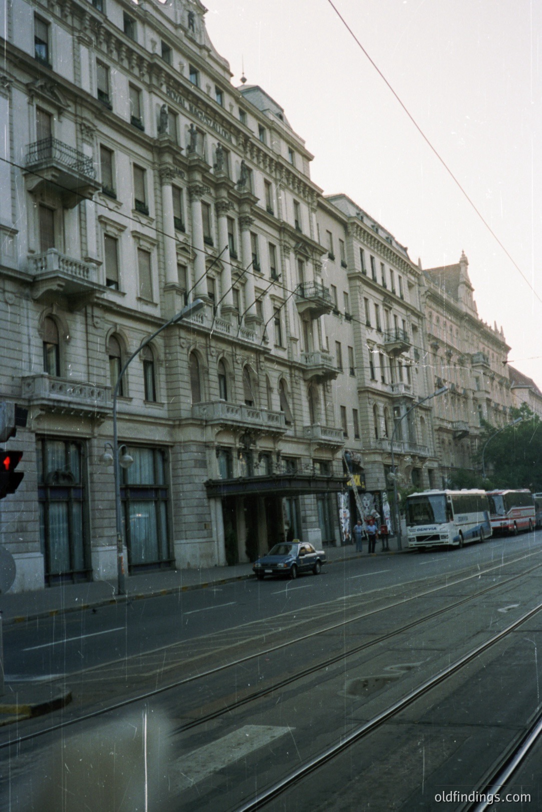 A wide, multistory building with ornate balconies lines a city street. Tram tracks run along the asphalt road. A bus and car are visible amidst pedestrians. Architectural style suggests early 20th century. Likely European city. Photograph has a slightly dated, film aesthetic.