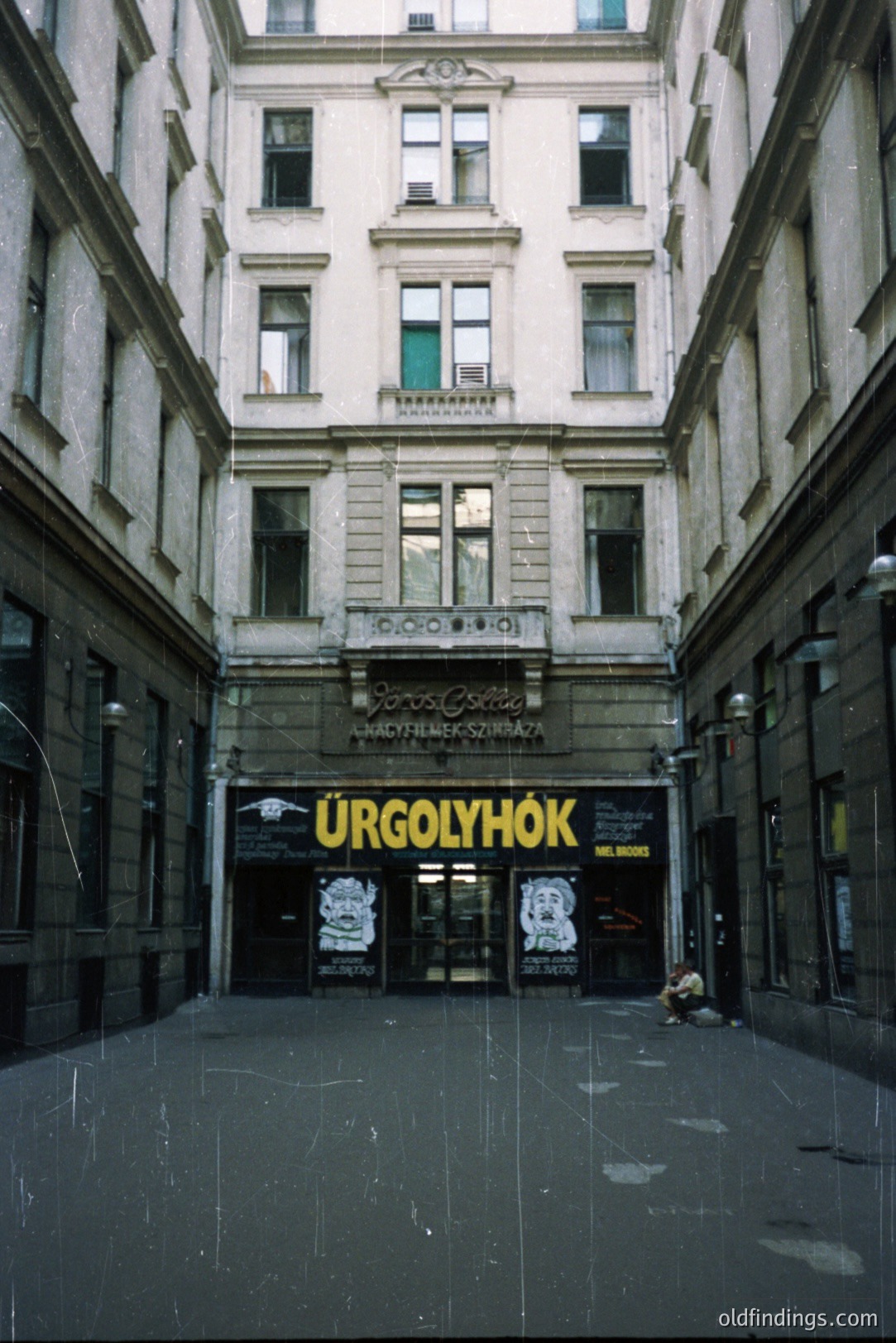 A long, narrow courtyard leads to a storefront entrance in a densely built urban area. The architecture features classical detailing, with decorative facades and window frames. "Ürgołyhók" signage is visible, suggesting a retail establishment. Likely Budapest, Hungary. Late 20th century photographic style.