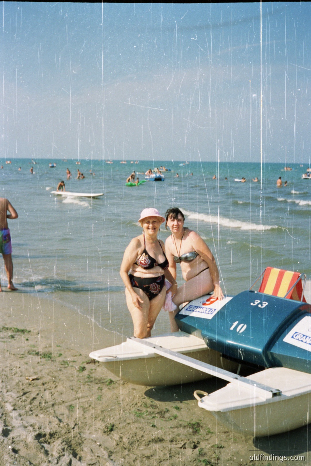 Two women in vintage swimsuits stand beside a small, numbered hydrofoil boat on a sandy beach. The scene includes a wide expanse of ocean and a bustling beach populated with people and recreational watercraft. The photo's fading and scratches suggest it's a snapshot from the 1970s or 80s.