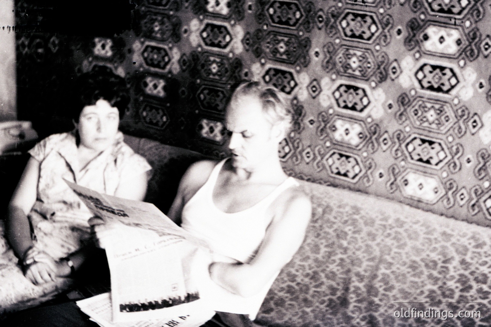 A man engrossed in reading a newspaper, seated on a patterned sofa. A woman is partially visible, seated behind him, appearing to observe. Interior scene with ornate, dark wallpaper and a distinctive, layered decorative cushion. Likely a domestic setting, 1940s-1960s.