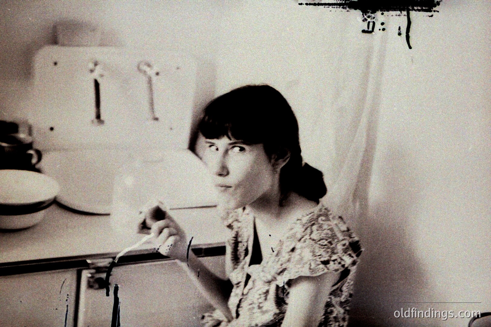 A young woman in a patterned blouse stands in a kitchen, pouring a liquid from a white pitcher. Mid-century modern kitchen features tiled walls and cabinetry. Likely a domestic scene; suggestive of 1960s or 1970s lifestyle photography. Strong composition for design or archival use.