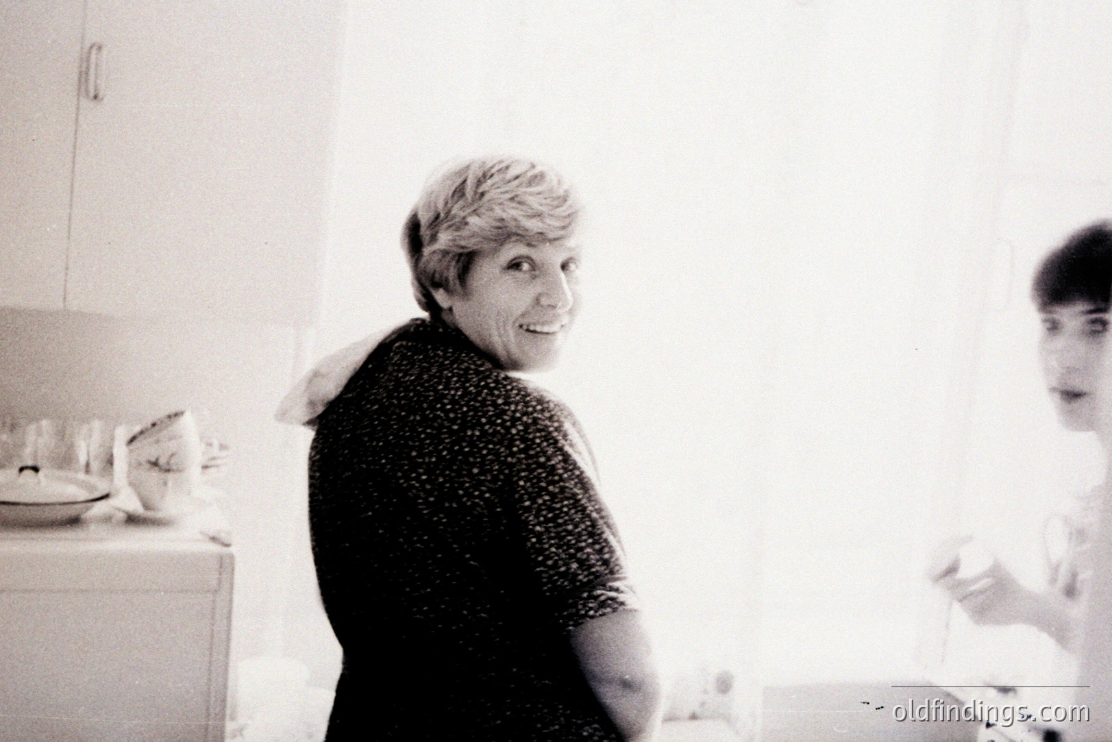 A smiling woman in a floral-patterned top looks back over her shoulder, while a younger person stands in profile near a kitchen counter. Domestic setting with dishes and glassware visible. Likely a snapshot from the 1970s, capturing candid family life. Simple, relatable, and potentially valuable for nostalgic stock imagery.