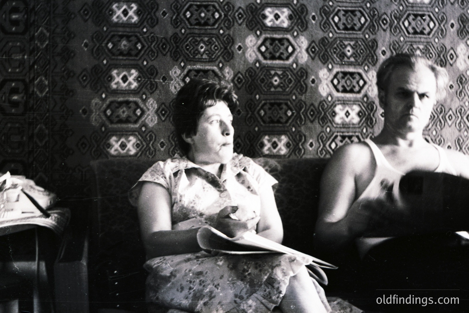 A woman seated on a sofa, holding a document, is framed by a vibrant, patterned wallpaper. A man sits beside her, appearing contemplative. Likely a domestic scene, possibly 1970s Eastern Europe. Strong visual texture & graphic design elements.