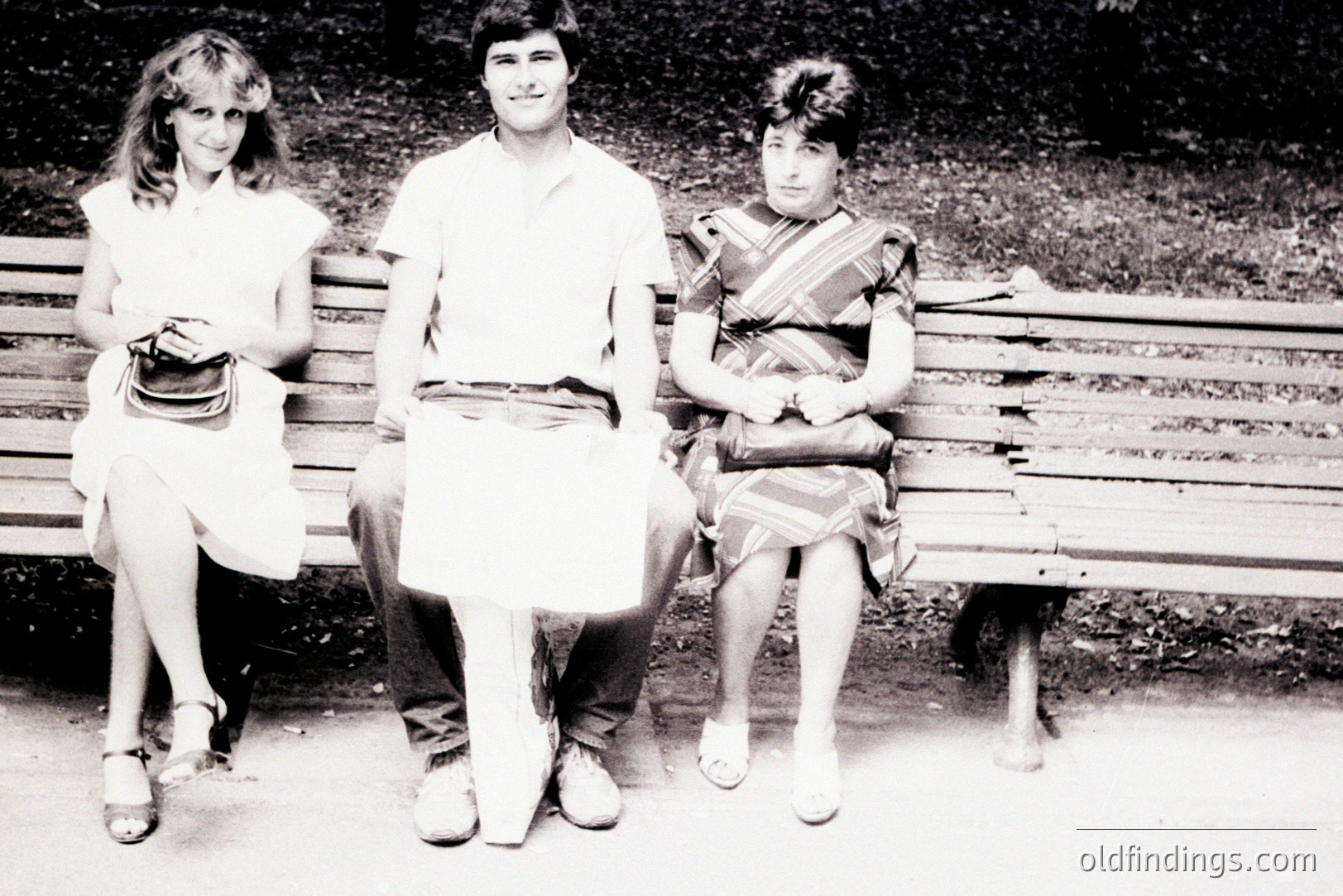 Three young people sit on a weathered park bench. The man, centered, holds a sheet of paper; the two women are on either side of him. Likely a candid, posed portrait. Fashion suggests the 1970s, with platform shoes & layered hairstyles. Location appears to be Eastern Europe. A classic, nostalgic scene.