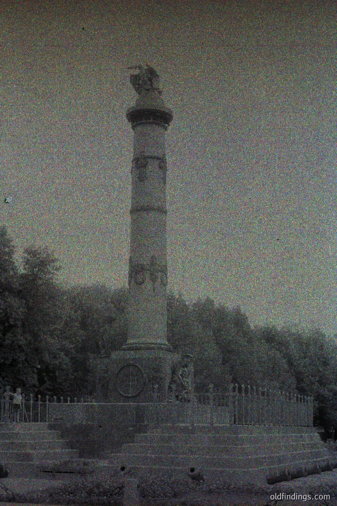 Monumental column with visible relief carvings rises from a stone base and ironwork fence, set against a backdrop of dense foliage. Likely a commemorative structure, the style evokes classical influences. Estimated time period: late 19th or early 20th century. Possible archival value for architectural or historical research.