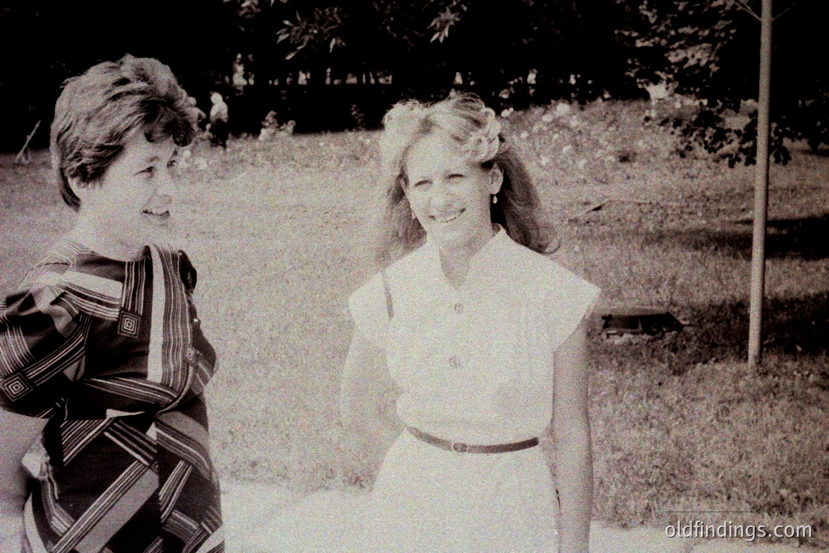 Two young women in casual summer attire, standing and smiling towards each other in a grassy outdoor setting. The woman on the left wears a striped, patterned dress. The woman on the right has a floral headband. Likely a candid snapshot, reflecting 1970s fashion and style. Appears to be a personal photo, potentially family archive material.