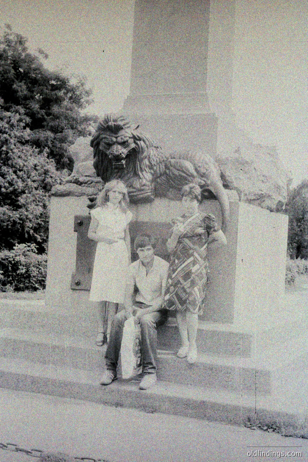 A black and white photo shows three people posed beside an ornate stone lion statue. Two teens sit on the steps, one girl stands adjacent. Likely a public park or garden setting, evoking a 1970s aesthetic. The statue's design suggests European influence, possibly Eastern European. Documenting a moment in time, capturing youthful style and a public space.