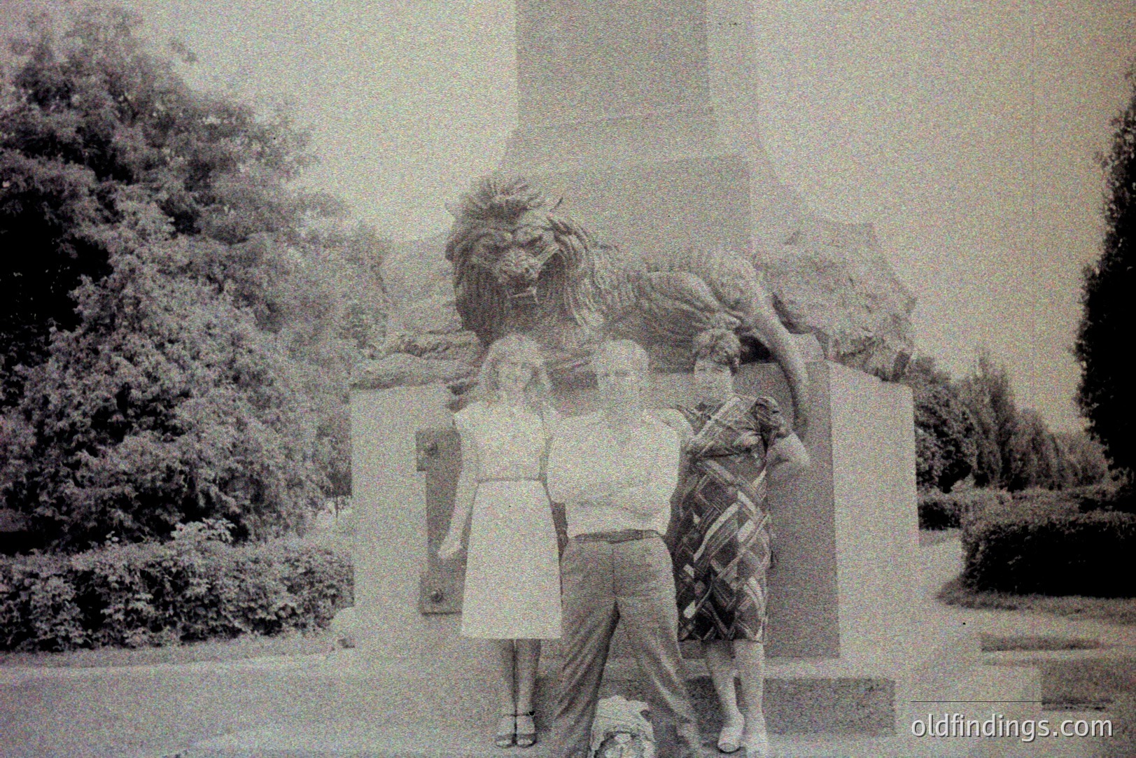 Three young people pose in front of a monumental stone sculpture depicting a lion and female figure – possibly allegorical. Architectural style suggests European influence, potentially Eastern European. Likely mid-1970s, based on clothing styles. The scene evokes a public space or park setting. Image has archival value.