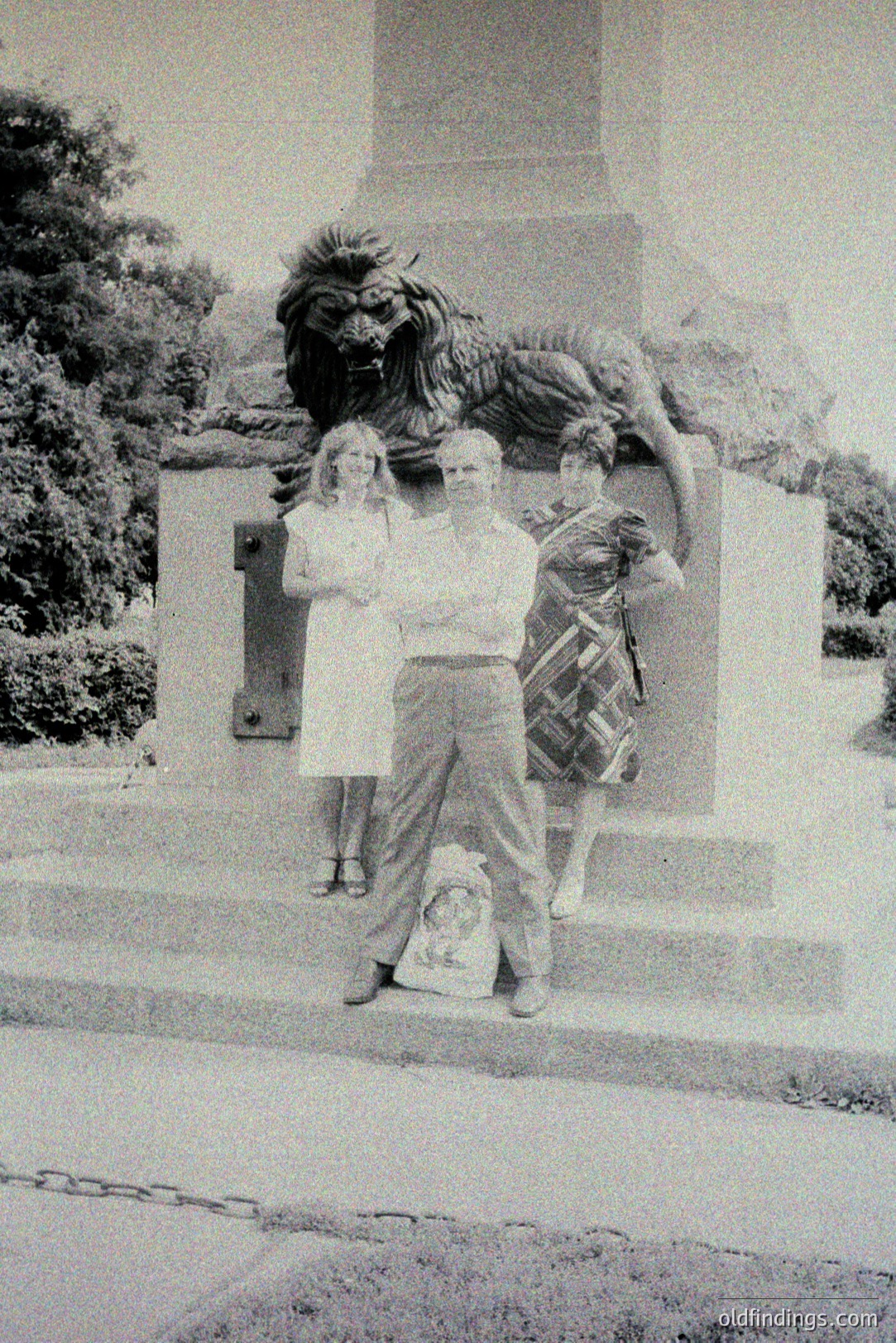 Three people pose before a monumental bronze statue depicting a lion, potentially a symbolic or allegorical representation. The individuals are dressed in 1970s-style clothing. A small portrait rests at the statue's base. Likely a travel snapshot, evoking a sense of mid-century tourism and public art.