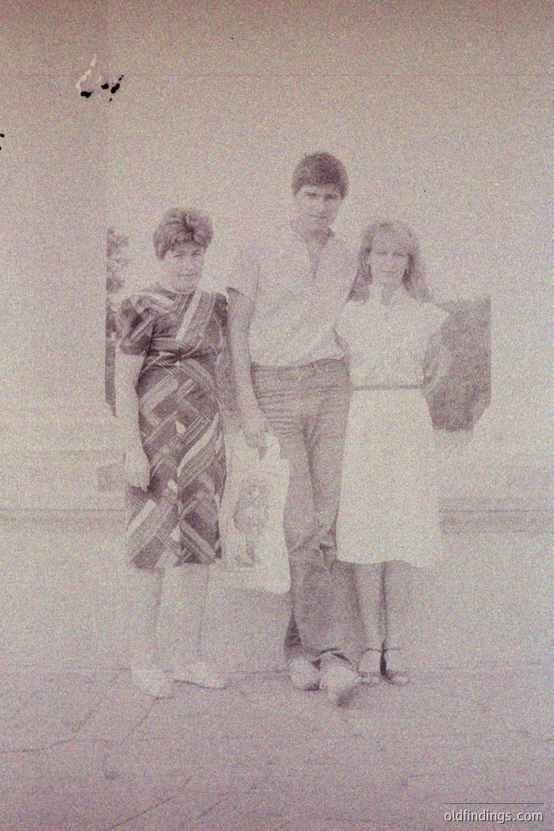 Three young individuals stand in a casual, outdoor portrait. The girl on the left wears a patterned dress, the boy in a button-down shirt and bell-bottom jeans, while the girl on the right wears a simple, knee-length dress. Clothing suggests a 1970s timeframe. A slightly blurred background hints at an open space. Likely a family snapshot.