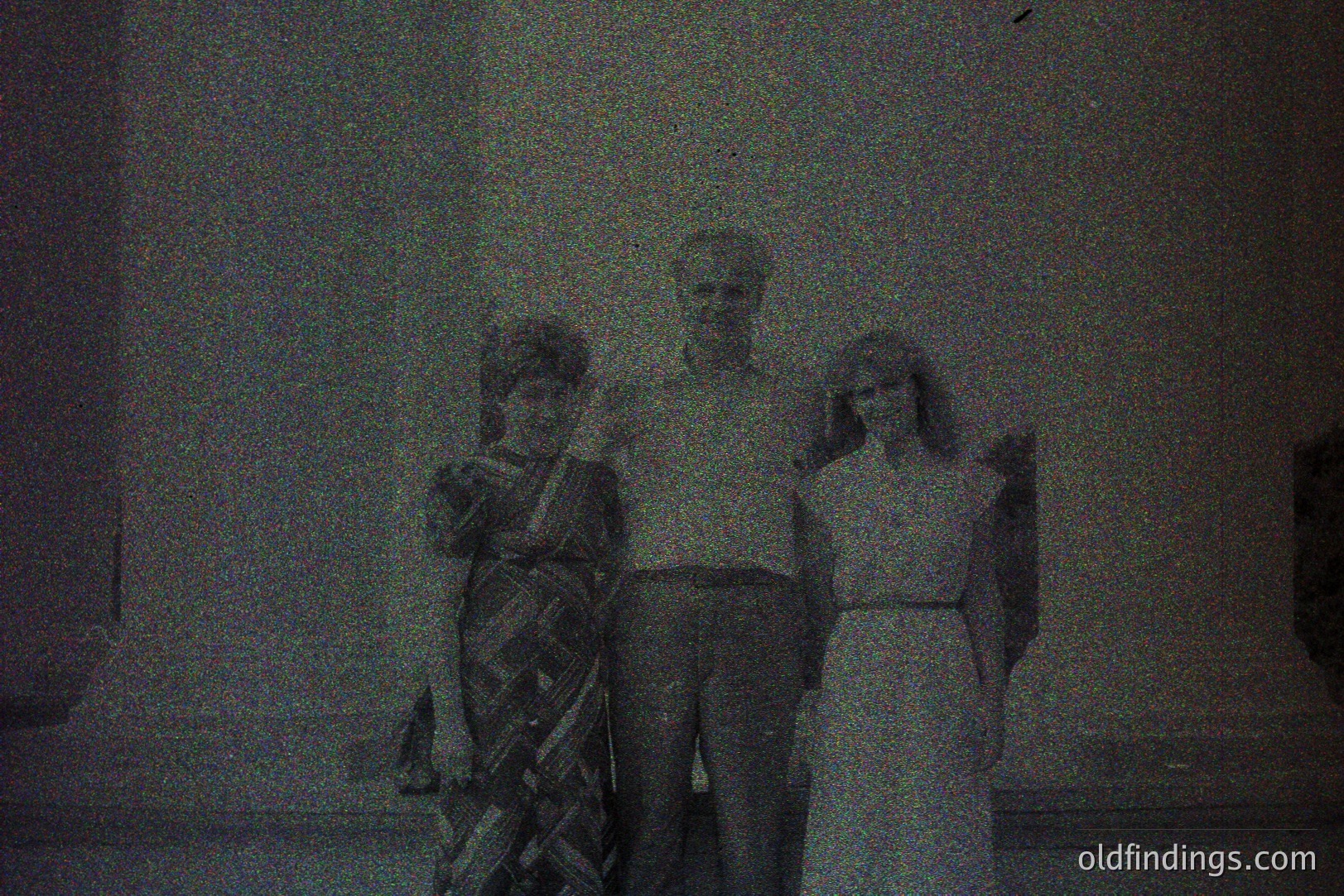 Three people stand posing outdoors, likely in front of a building. The woman on the left wears a patterned dress, the man is center, and the woman on the right wears a long, simple white dress. The grainy quality suggests a vintage print, possibly 1970s. Architectural detail visible hints at Mediterranean design.