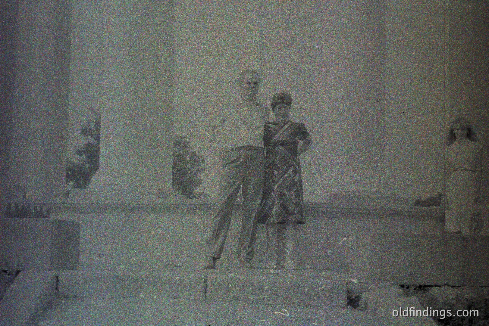 A grainy black and white image depicts three figures on tiered steps. Two adults, a man in a short-sleeved shirt and a woman in a patterned dress, stand close together. A third person, partially visible, stands further back. Likely a public space. Appears to be a candid family portrait.