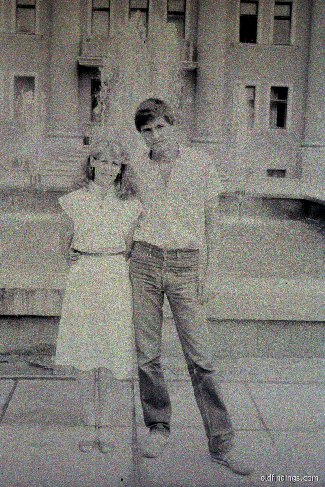 A young couple stands outside a classical building featuring a balcony with statues. The woman wears a short, ruffled dress; the man, a button-down shirt and flared jeans. Likely 1970s fashion and architecture. Location unclear. Evokes a sense of youthful nostalgia. Potential for design/vintage marketing.