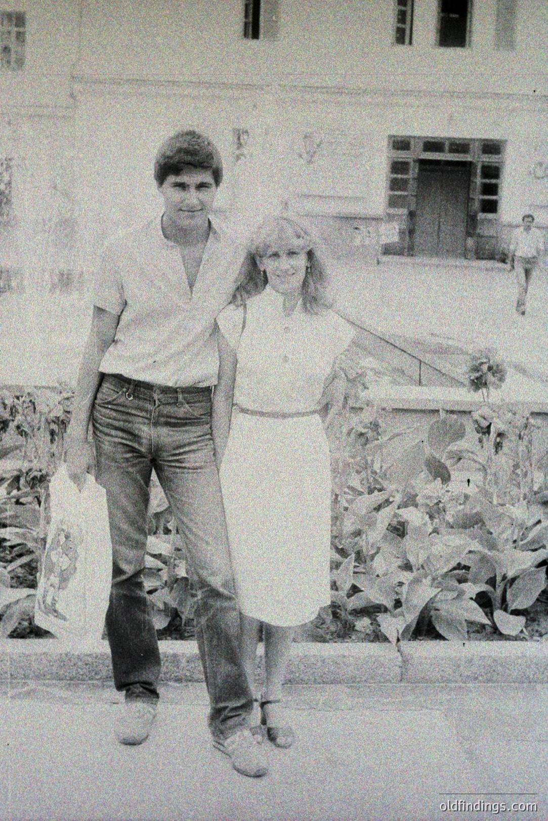 A couple strolls along a paved walkway in front of a stately building featuring arched windows and ornate detailing. The man wears a short-sleeved shirt and flared jeans, while the woman wears a simple dress. A shopping bag is visible. Likely 1970s.