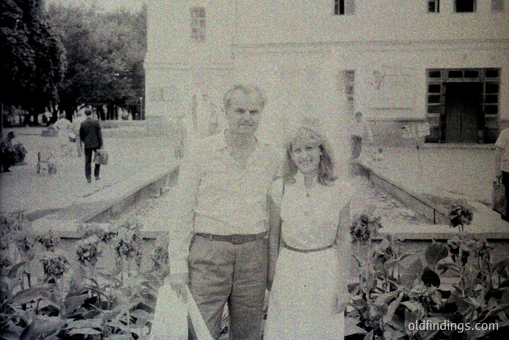 A black & white snapshot depicts a couple posing formally in front of a large, classically styled building with a prominent portico. The architecture suggests a European estate or public building. Man in slacks & button-down; woman in a long dress. A figure walks in the background. Likely 1970s.
