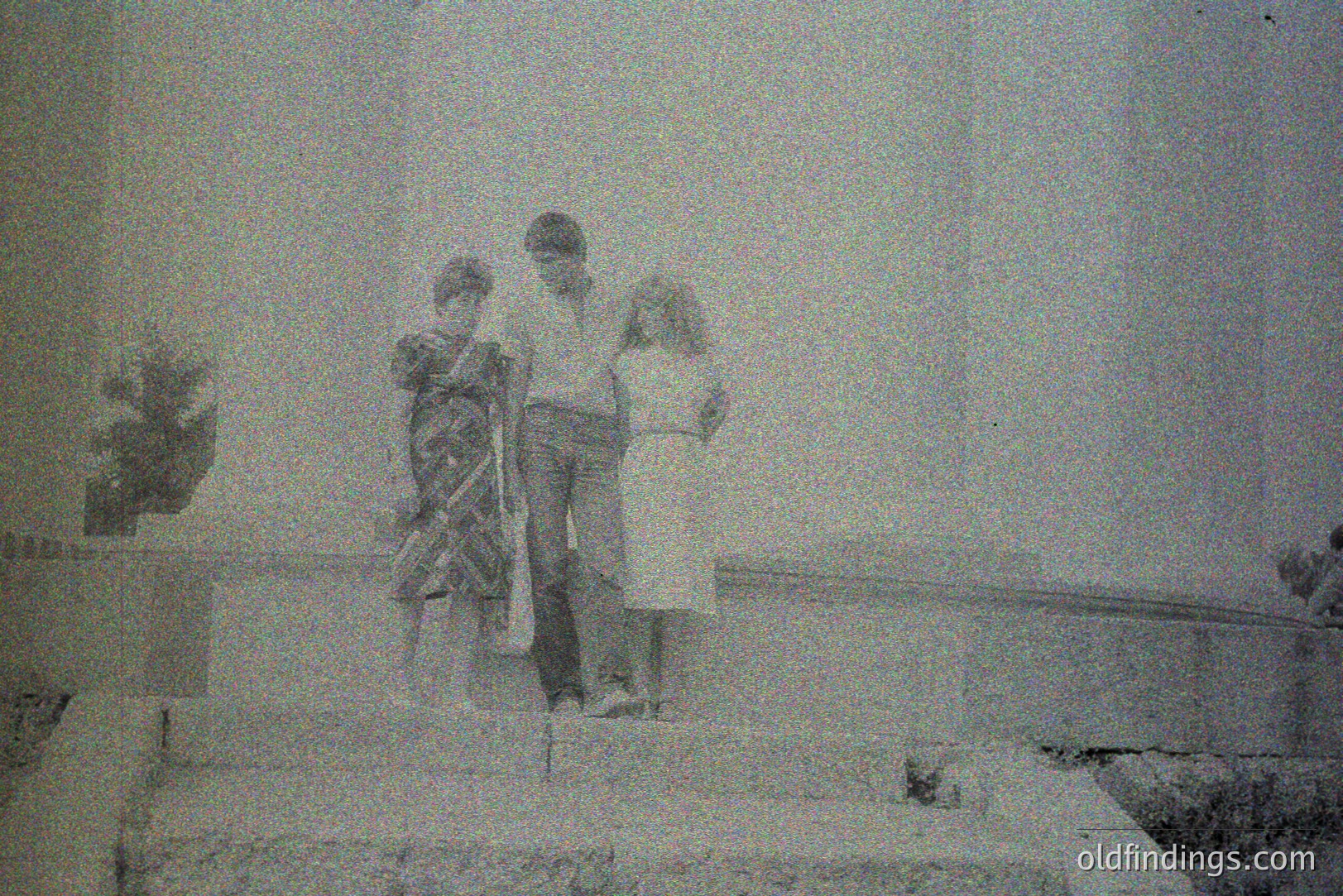 Three children stand on a stone ledge or staircase in what appears to be an architectural setting, possibly a courtyard or public space. The child on the left wears a patterned dress, while the others are in simple attire. Grainy black and white image suggests a candid moment, likely 1960s-1970s. Likely an amateur photograph with potential for historical family archives.