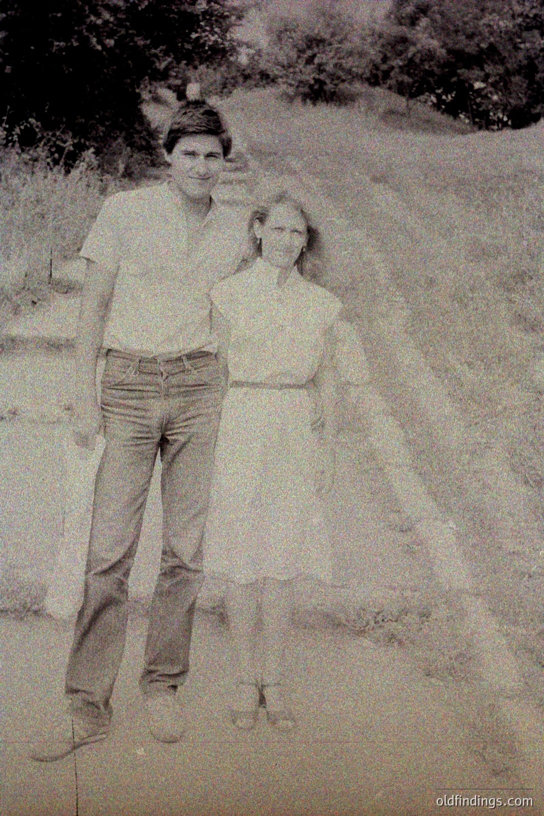 A young man and girl stand outdoors on a gravel path. The man wears a short-sleeved shirt & jeans; the girl wears a sleeveless dress with a Peter Pan collar. Likely 1970s casual portrait with natural lighting and grainy texture. Rural setting suggests a vacation or family outing. Could be valuable for period design references.