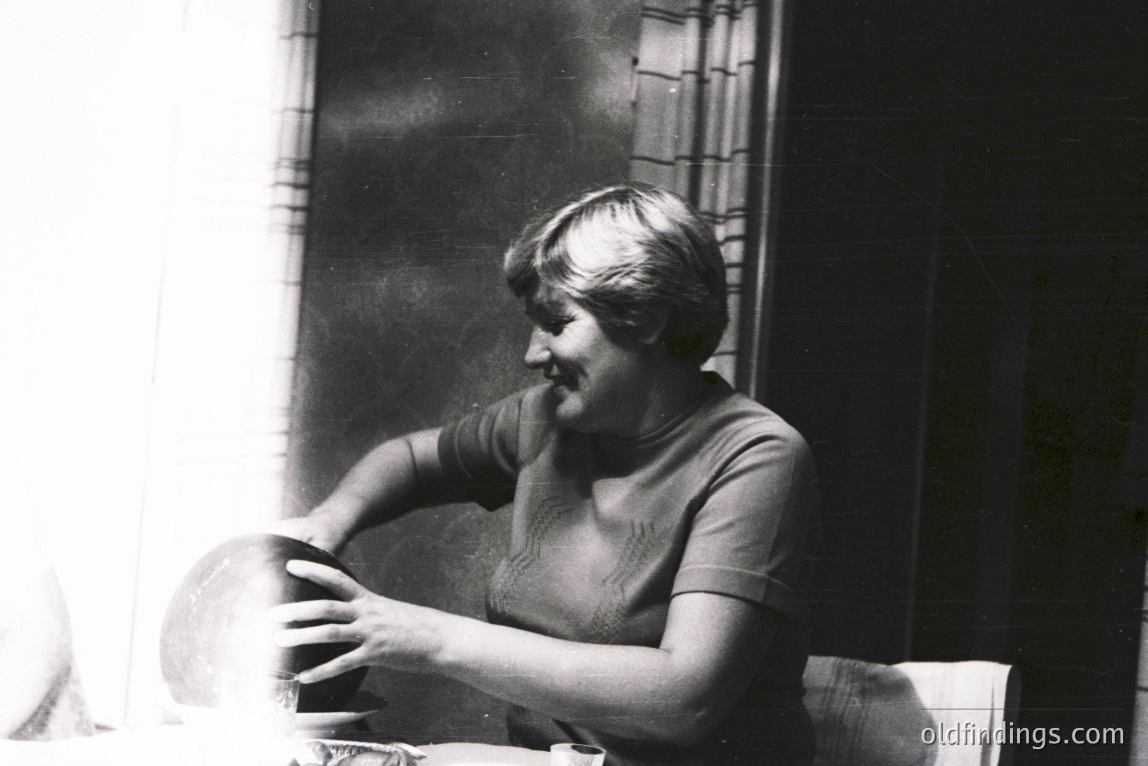 A woman in a textured knit top sits at a table, carefully cradling a ceramic bowl. The scene is brightly lit, creating strong contrast. Likely a domestic snapshot, possibly 1970s, capturing a moment of everyday life. Good reference for design or vintage advertising.