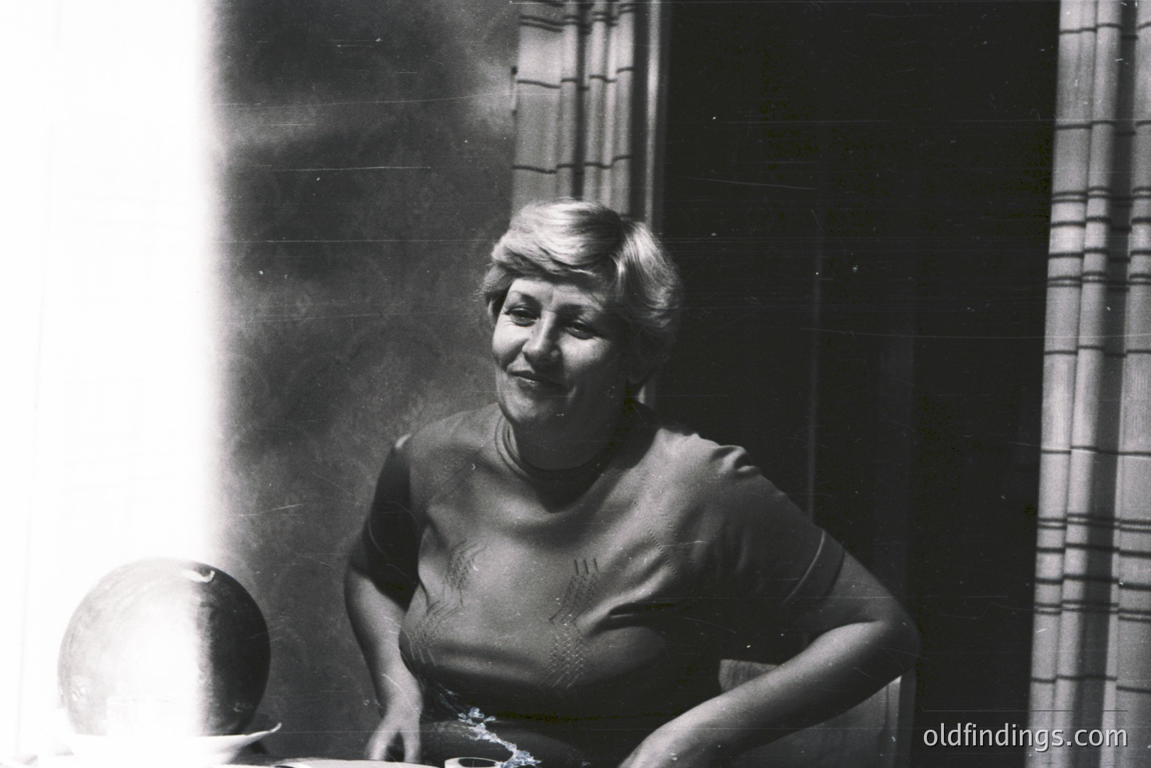 A smiling woman with a short, styled haircut sits at a table in a dimly-lit room. A sculpted head rests on the table. Drapery is visible behind her. Appears to be a candid portrait, possibly 1960s-1970s. Likely domestic setting.