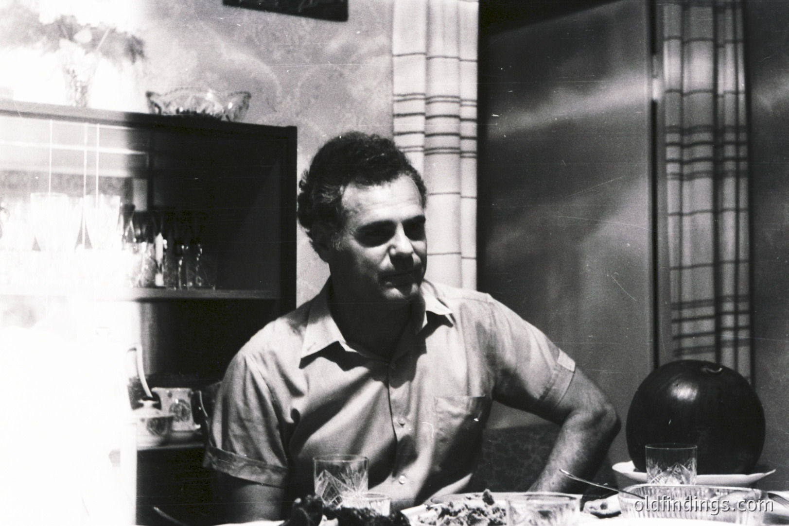 A seated man in a short-sleeved shirt appears to be at a table setting with glassware and a dark, round object. Interior scene shows patterned wallpaper and a cabinet with bottles. Likely a candid snapshot, possibly 1970s. Sharp focus, strong contrast.