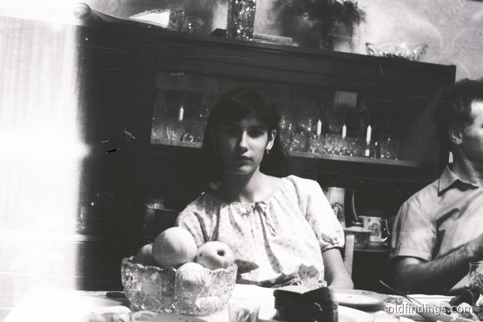 A young woman sits at a table, facing the camera, with a bowl of fruit beside her. A man is partially visible to the right. Interior scene with a large, ornate cabinet in the background. Likely a family gathering. Appears to be from the 1970s.