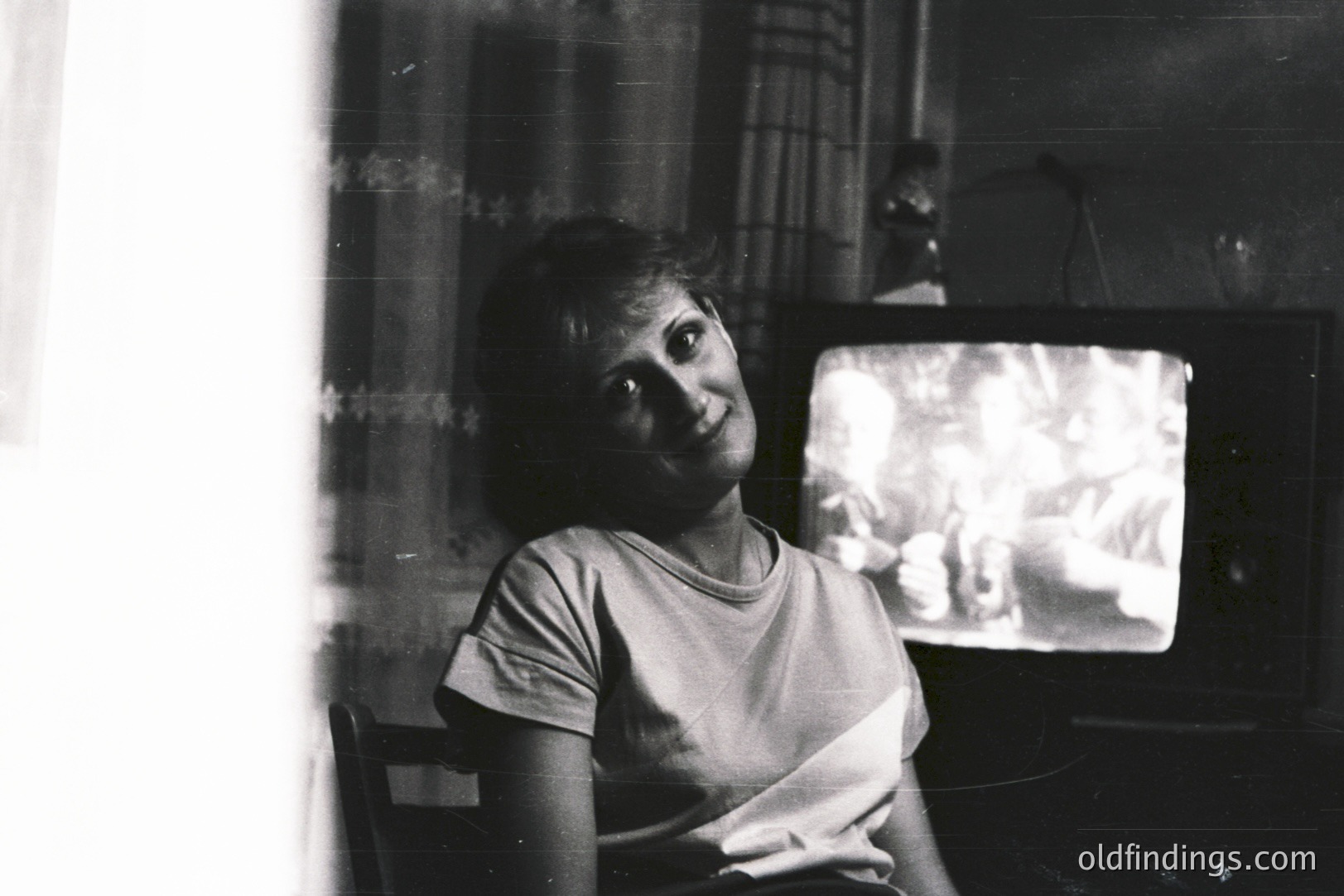 A young woman sits facing the camera, partially obscured by a bright window. She wears a graphic tee, seated near a vintage television displaying nature imagery. Likely a candid portrait from the 1970s, evoking a domestic scene and showcasing early television technology.